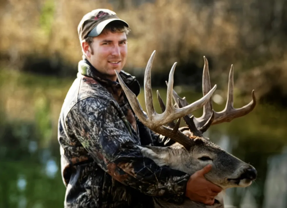 Darren Kalin posing outdoors, smiling, wearing a camouflage jacket and a brown hat, and holding up the head of a large whitetail deer buck with substantial antlers.